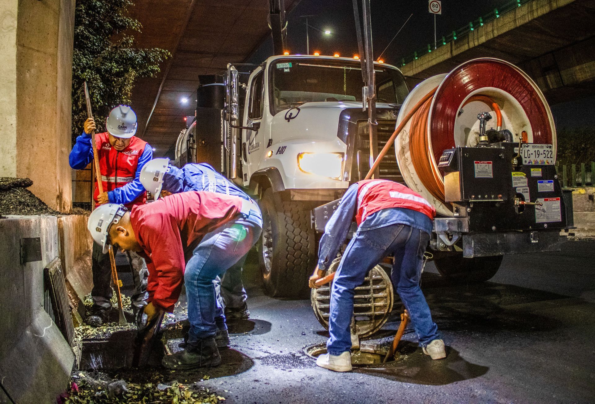 Utility workers repairing a manhole at night beside a truck, with bright work lights and wet pavement.