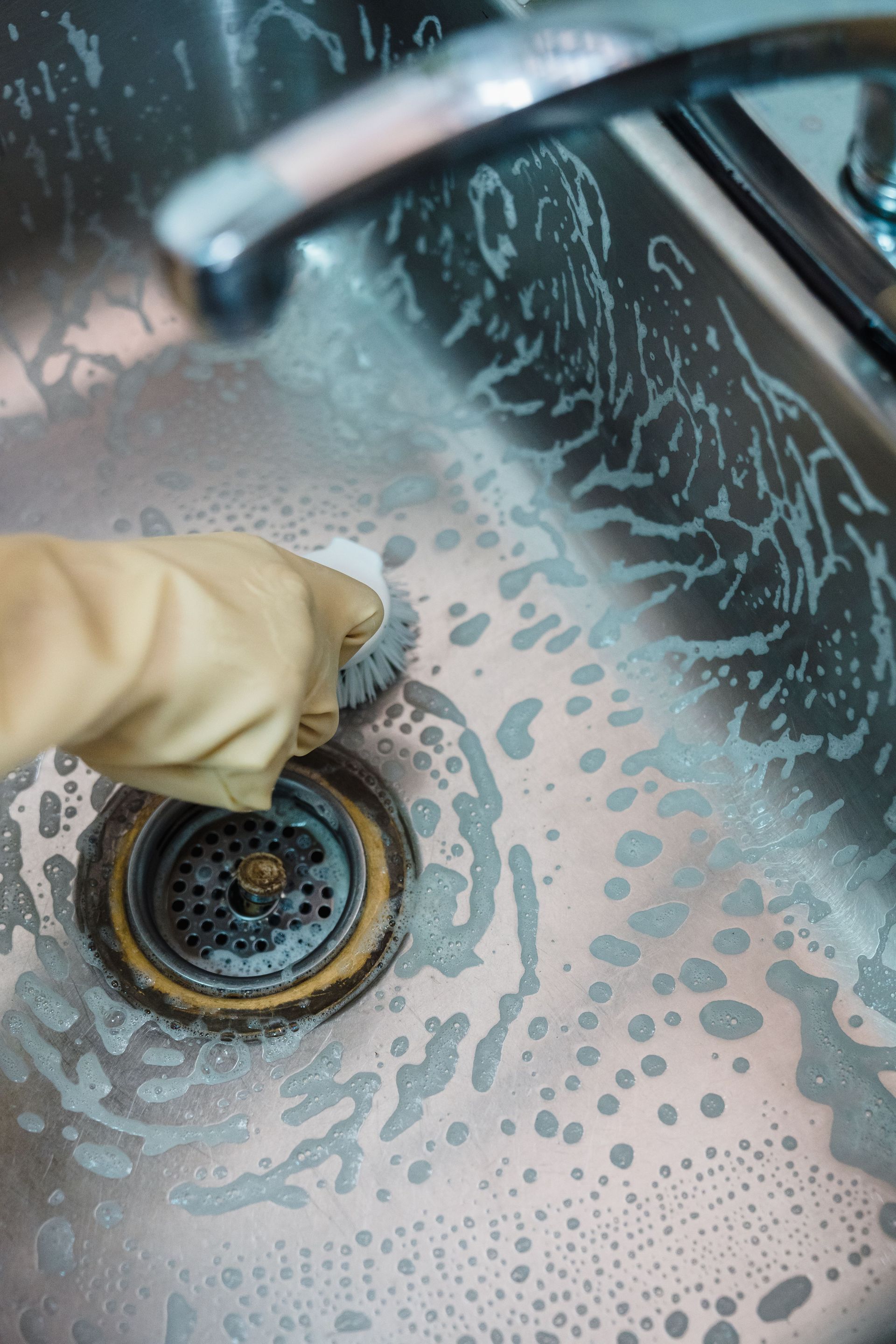 Gloved hand scrubbing a soapy stainless steel sink drain with a sponge