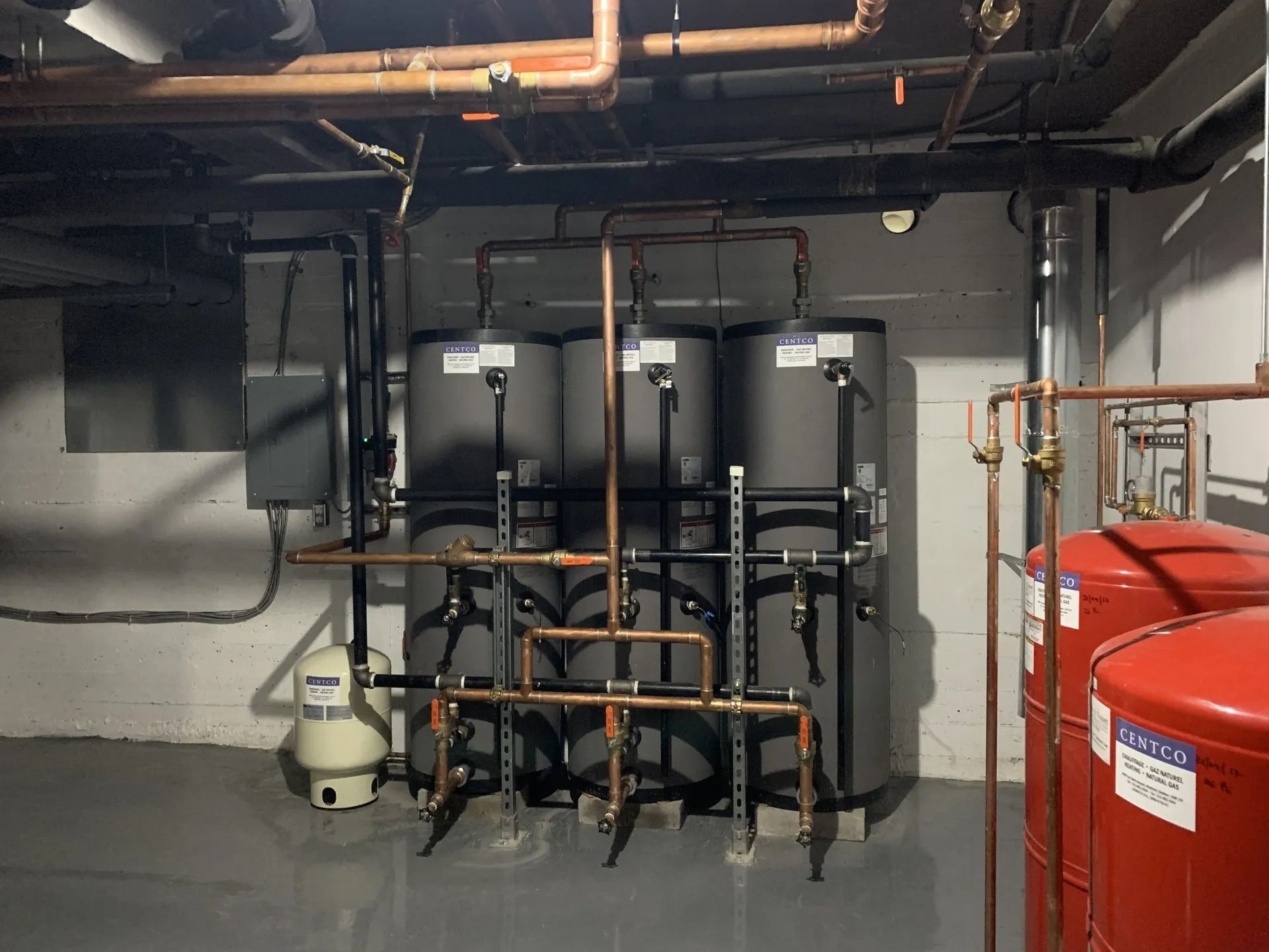 Local technique au sous-sol avec réservoirs d'eaux noires, tuyaux en cuivre et chaudières rouges sur un mur gris