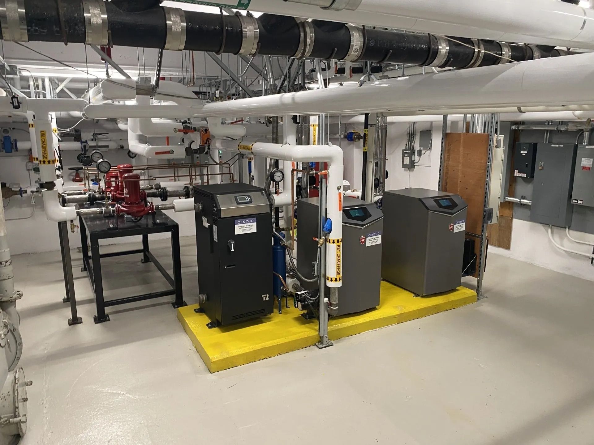 Mechanical room with pipes, pumps, and tanks on a yellow platform in a basement utility area