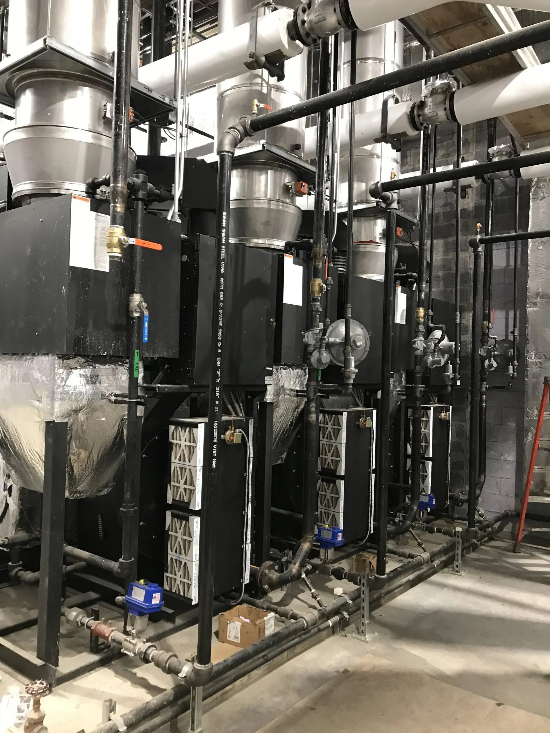 Industrial piping and black tanks in a mechanical room, with valves, valves, and metal ductwork.