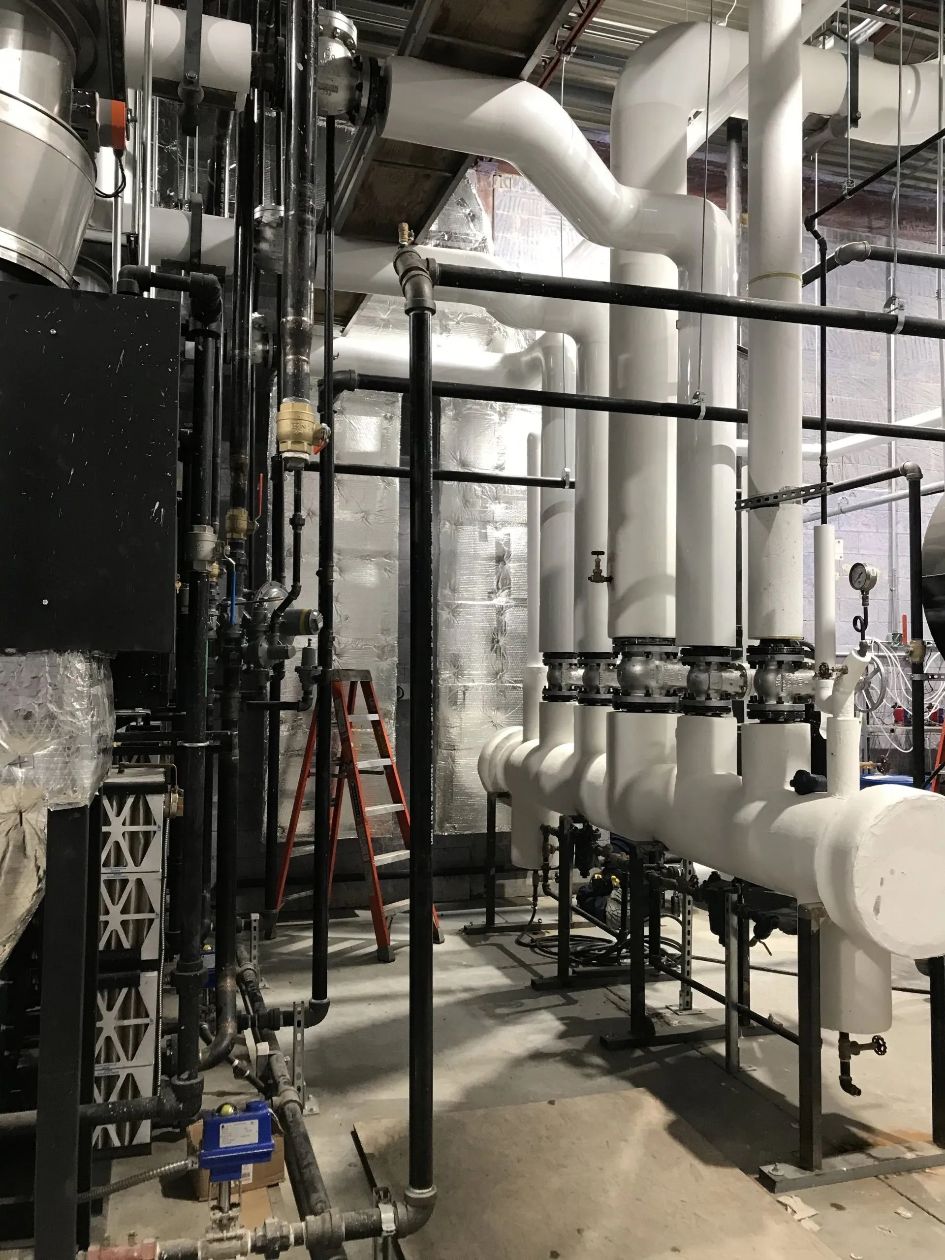 Industrial mechanical room with large white insulated pipes and valves, a red ladder, and metal piping supports