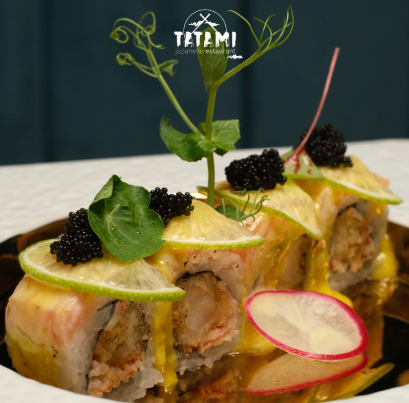 Uramaki guarniti con avocado, caviale nero e ravanello, serviti su un piatto bianco e accompagnati da erbe aromatiche.