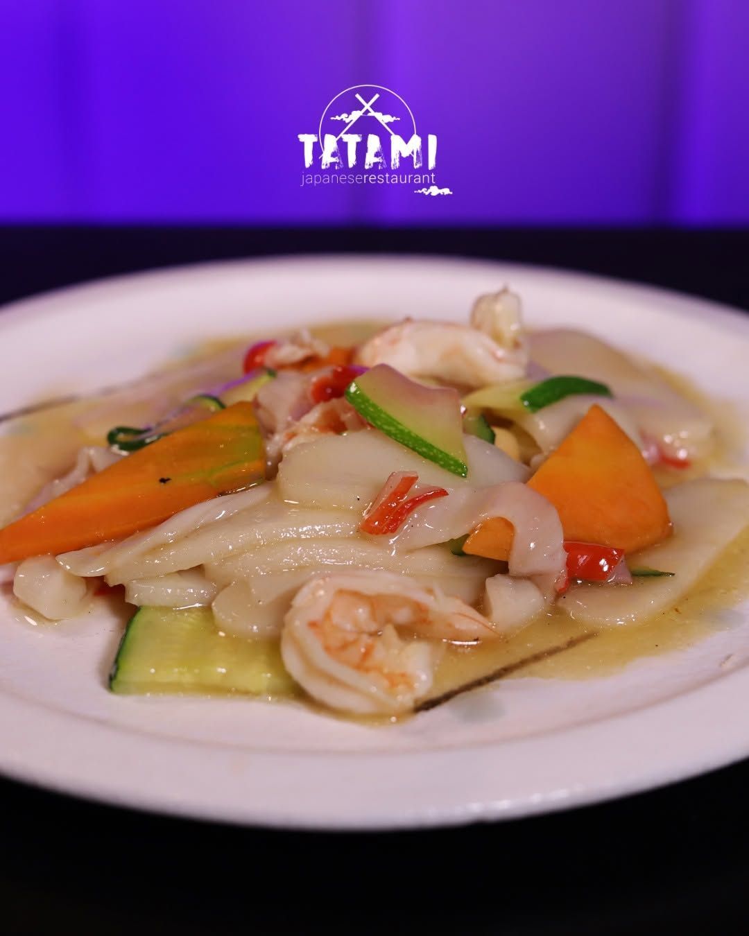 Piatto di gamberi colorati e verdure saltati in padella, serviti su un piatto bianco, con sullo sfondo il logo viola del ristorante