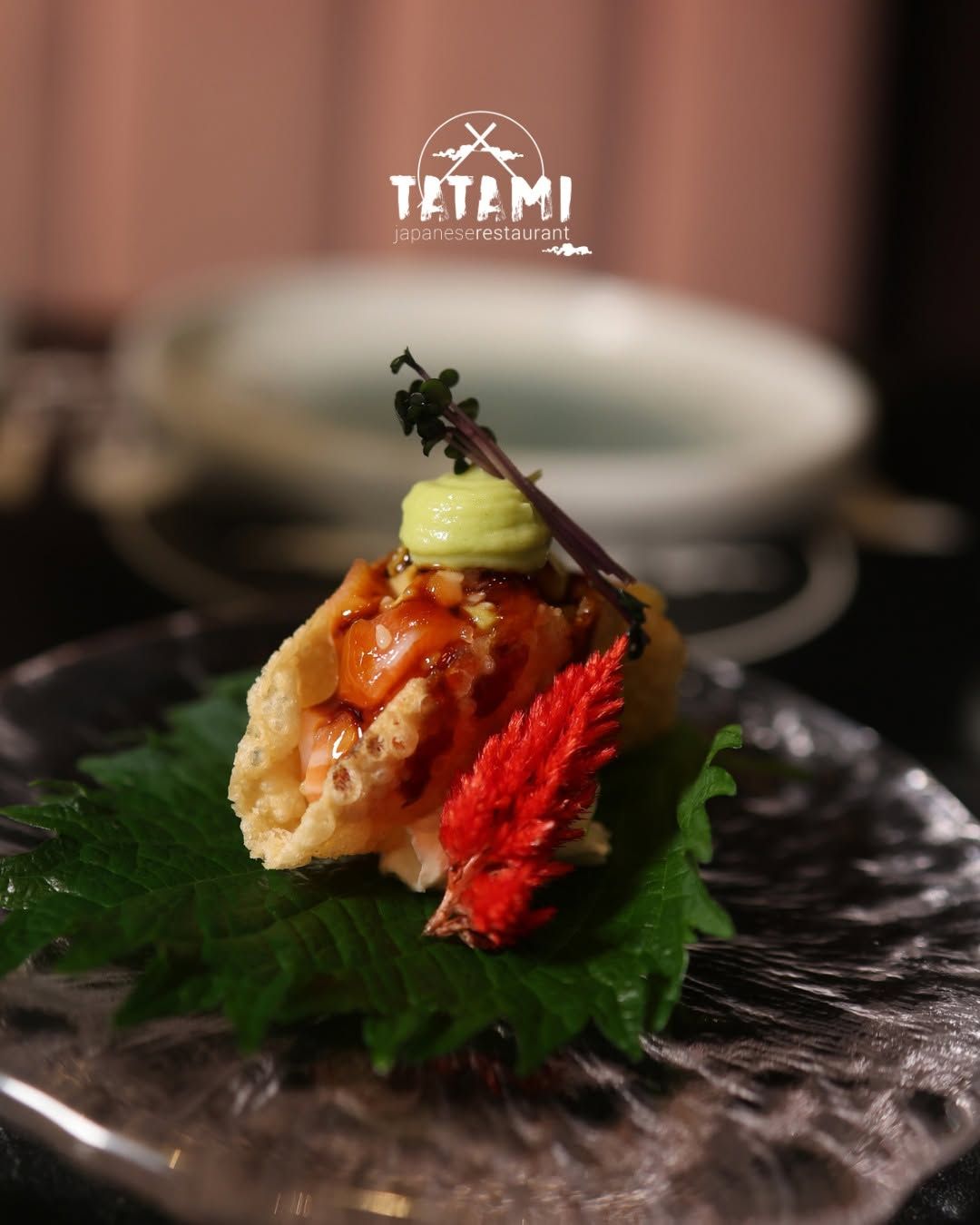 Rotolo di sushi gourmet guarnito con salsa, wasabi ed erbe aromatiche su un piatto nero, con un ristorante sfocato sullo sfondo
