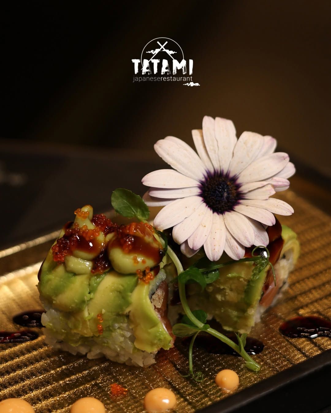 Due involtini di sushi guarniti con un fiore bianco, su un piatto dalla superficie ruvida con decorazioni di salsa a filo