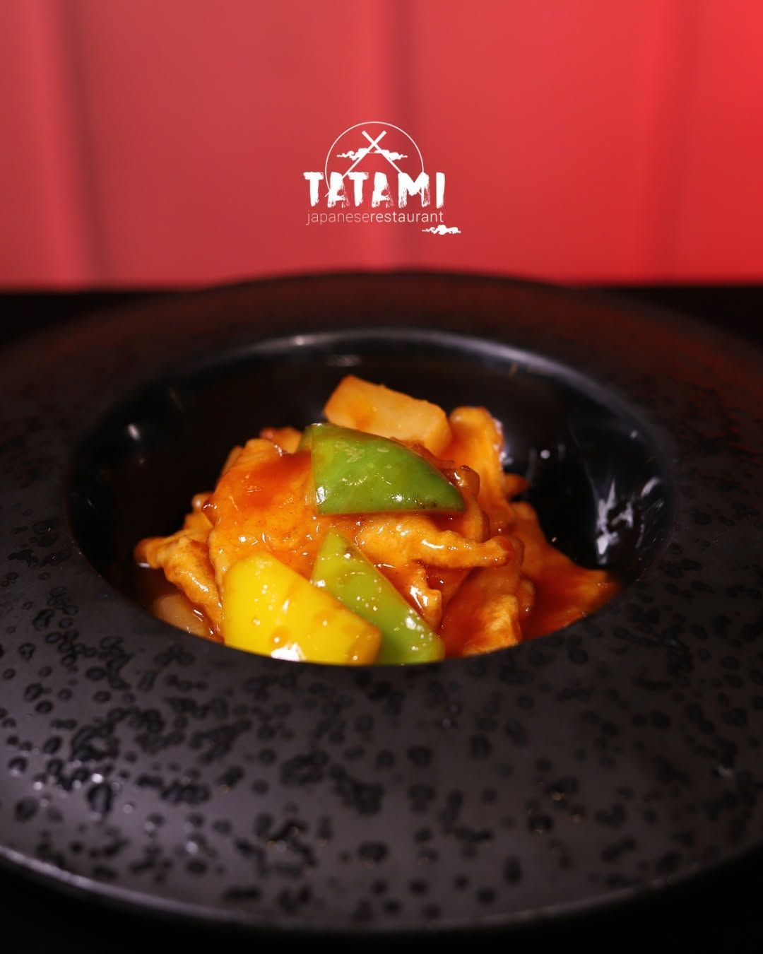 Ciotola nera dalla superficie ruvida con verdure condite con salsa all’arancia e peperone verde, su uno sfondo rosso da ristorante