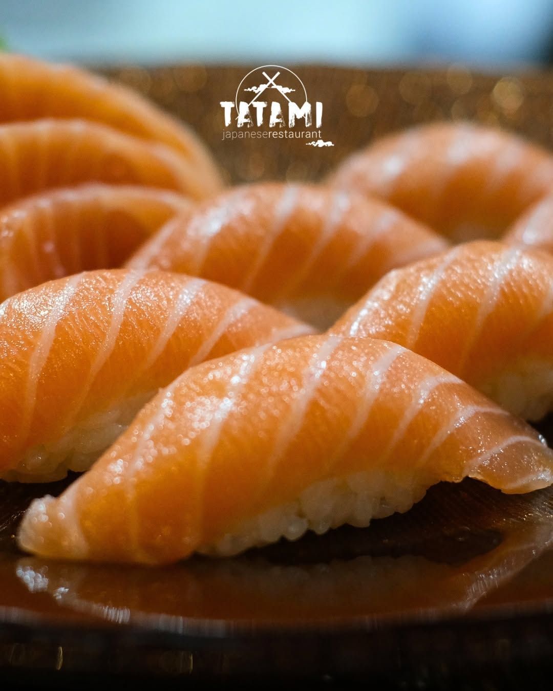 Primo piano di nigiri al salmone lucidi disposti su un piatto scuro, con lo sfondo sfocato di un ristorante