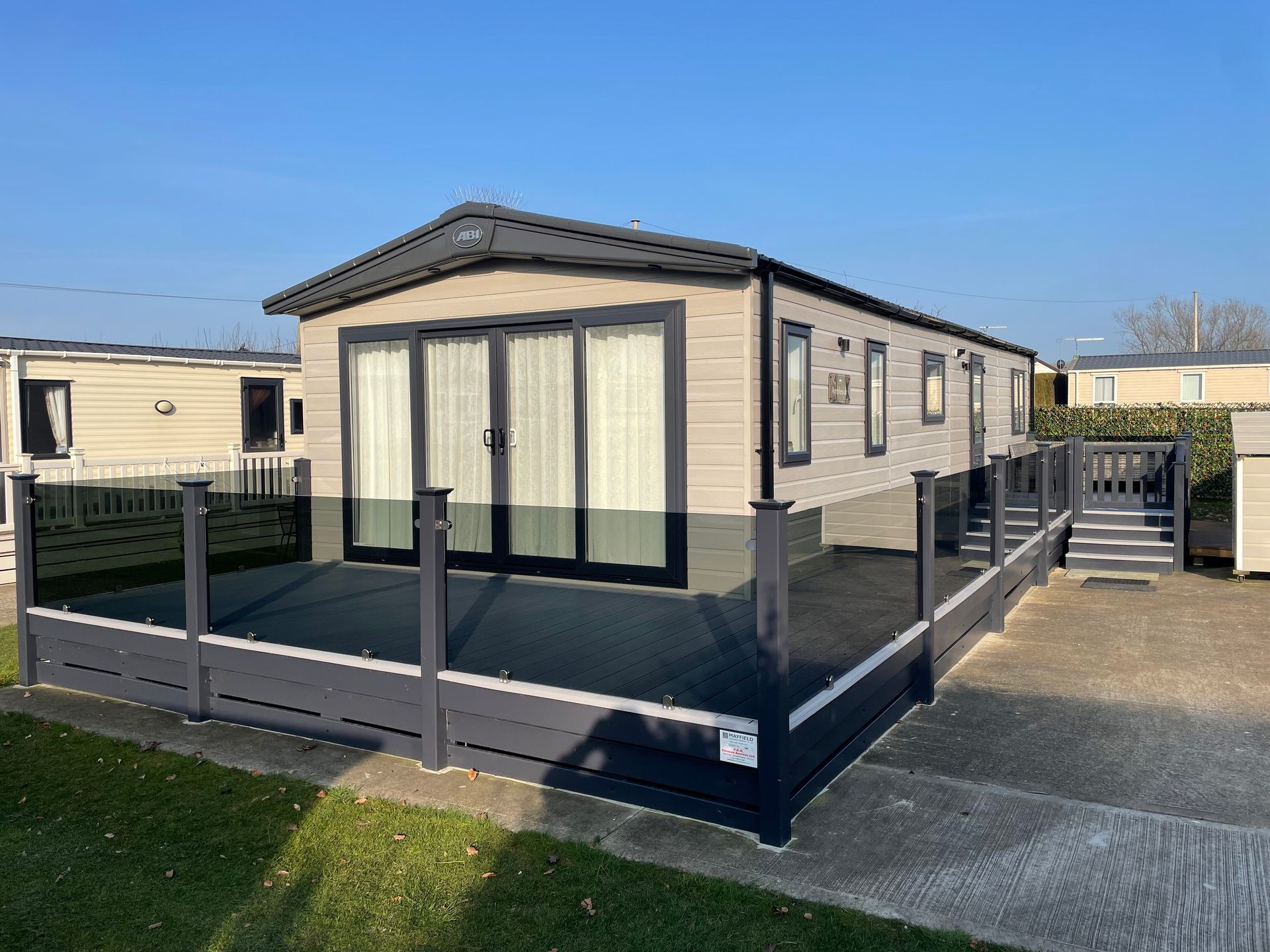 Mobile home with large deck, gray exterior, sliding glass doors, blue sky.