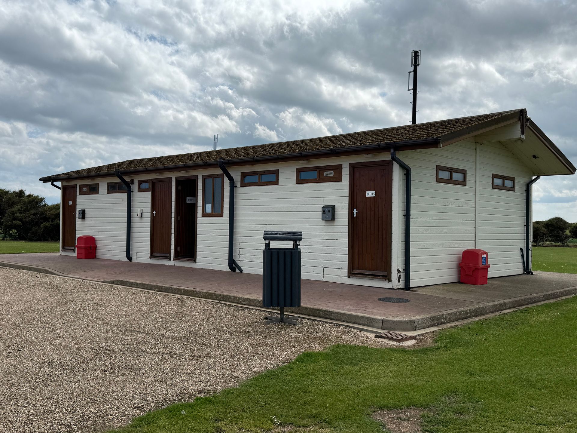 White public restroom with brown doors and roof on a grassy area under a cloudy sky.