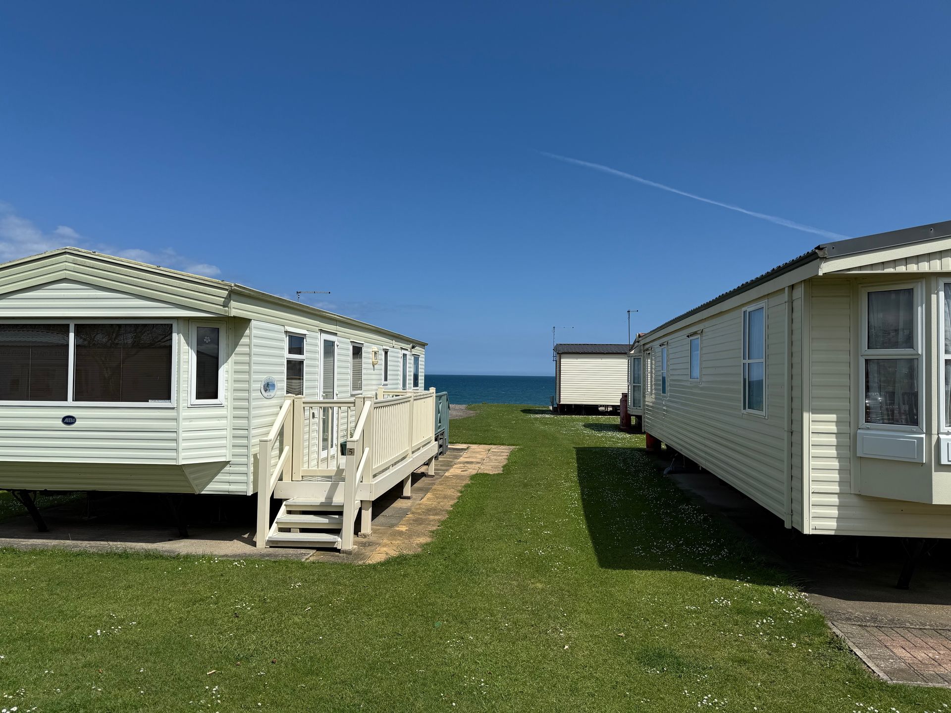 Two cream-colored caravans on a grassy lawn, with the ocean visible in the distance under a clear blue sky.