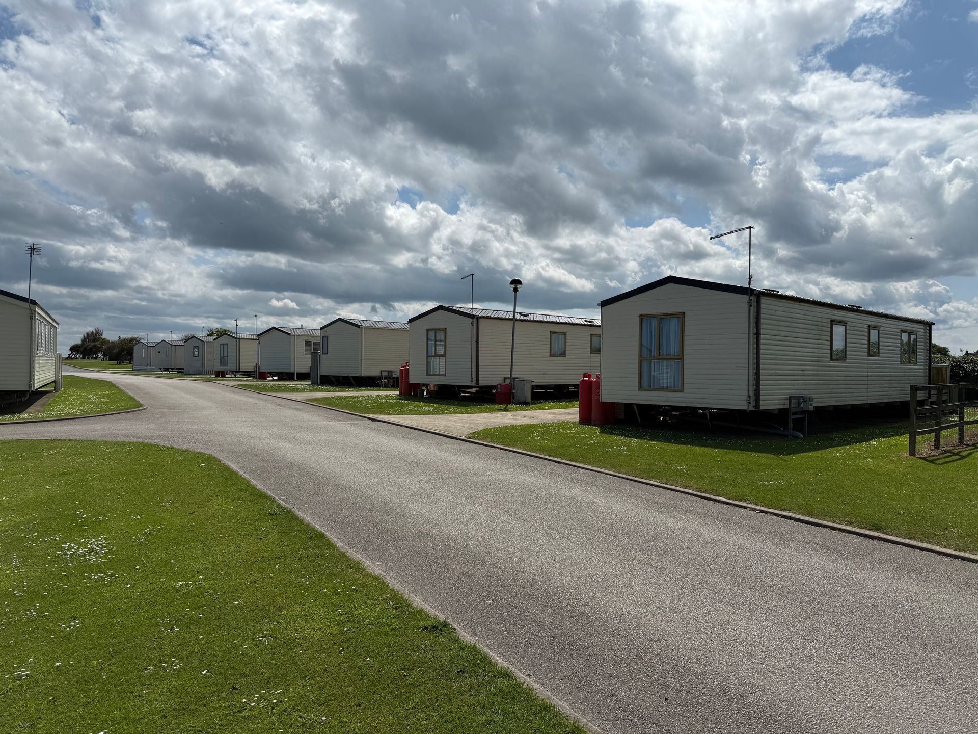Row of mobile homes on a paved road, grassy areas under a cloudy sky.