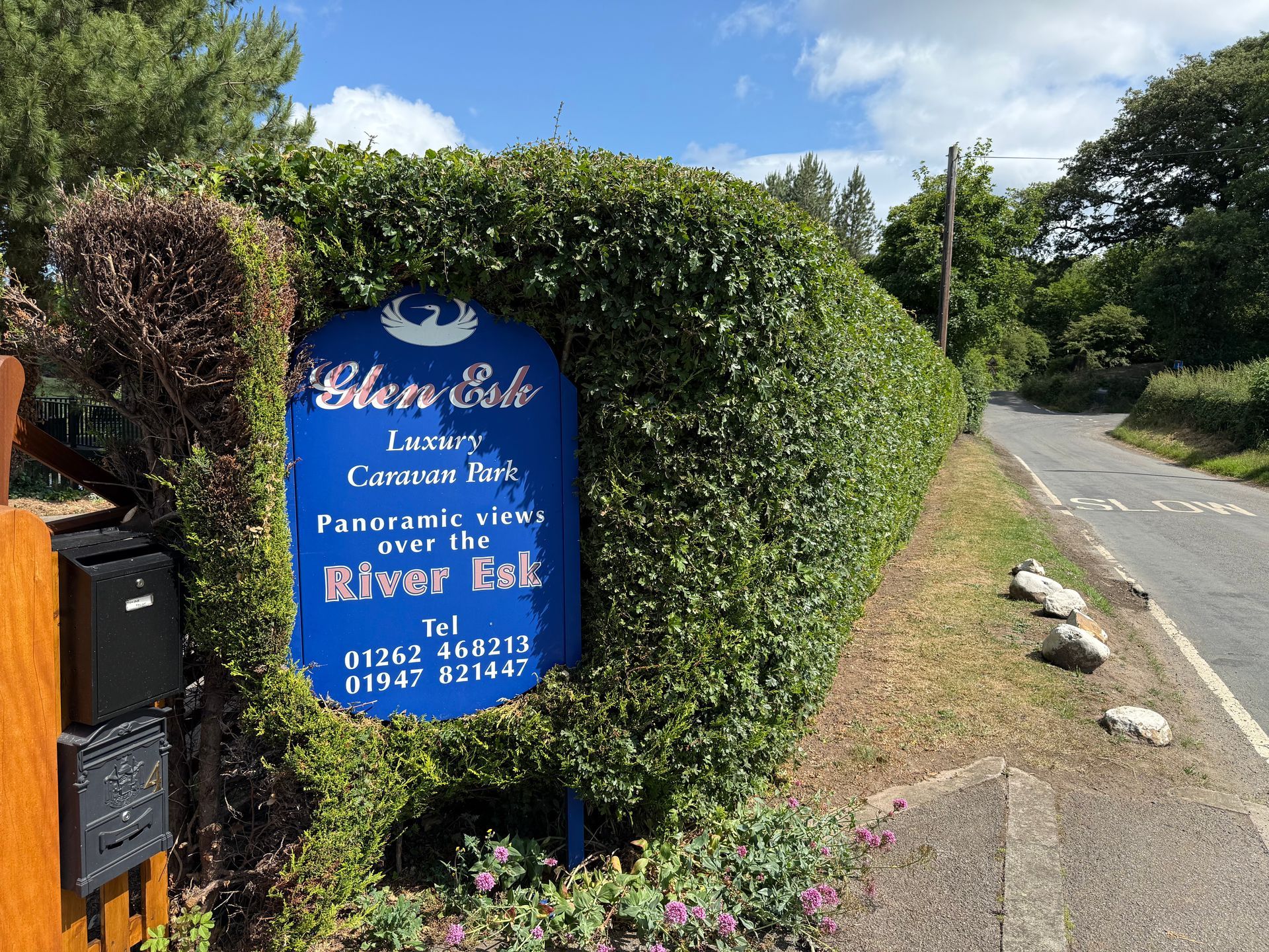 Sign for Glen Esk, luxury caravan park, overlooking River Esk. Blue sign with white text, framed by green hedge.