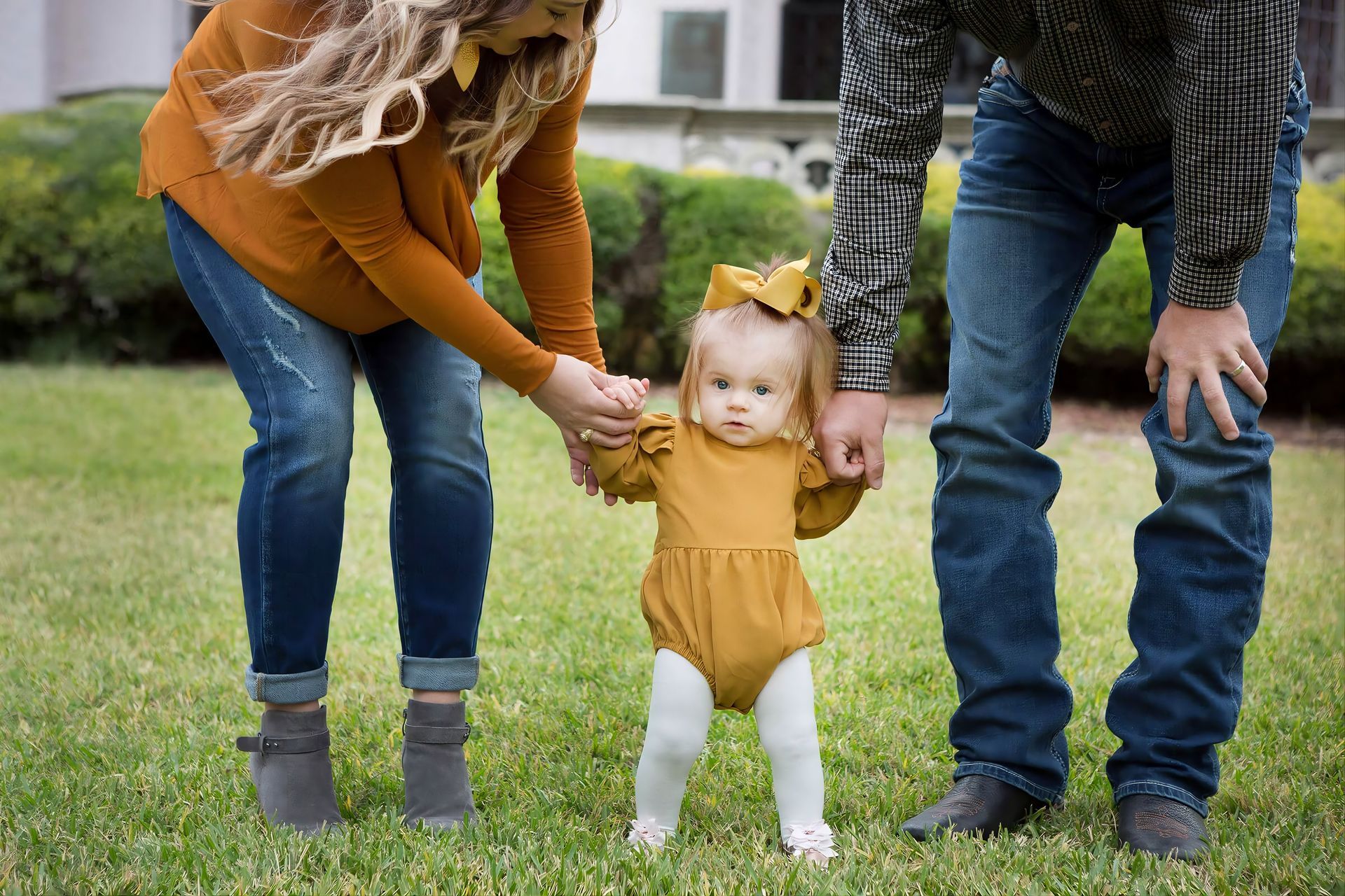 Two adults holding a toddler’s hands while she walks on grass in a park, wearing a mustard dress and white tights