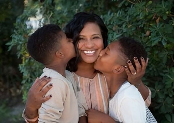Smiling person receiving kisses on both cheeks in front of leafy greenery