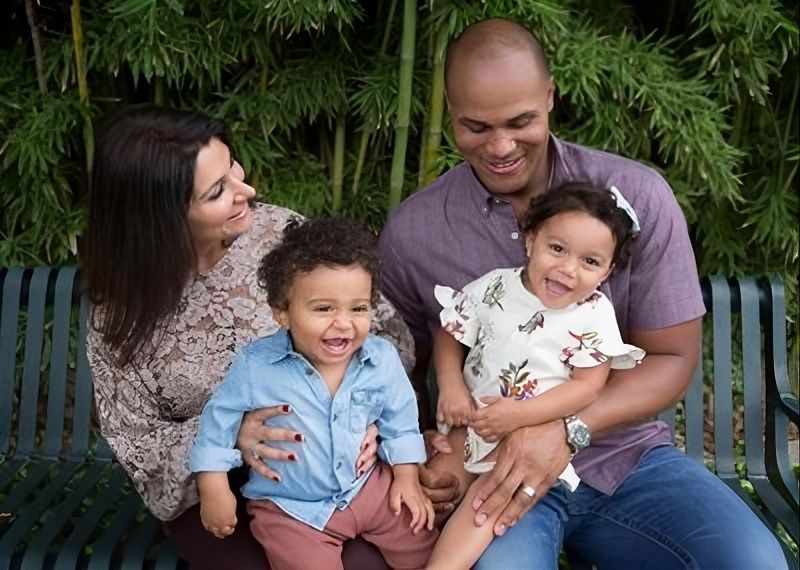 Family of four sitting together outdoors, smiling against a leafy green backdrop.