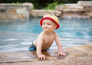 A toddler wearing a red and yellow hat smiles while leaning on the edge of a swimming pool.