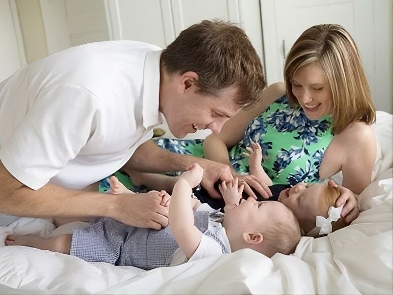 Family playfully interacting with a baby on a bed, smiling and leaning close together