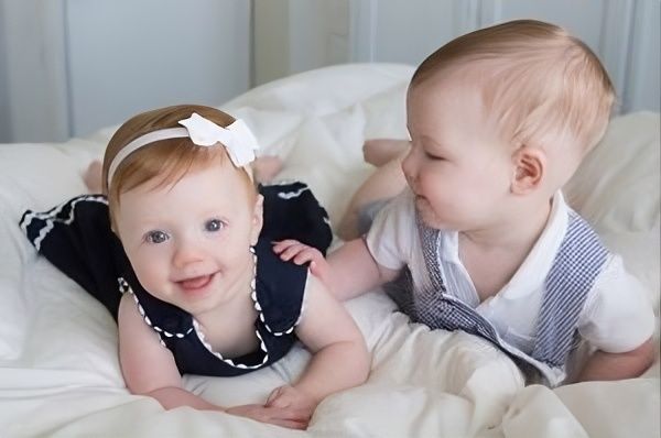 Two babies smiling on a white bed, one reaching toward the other.