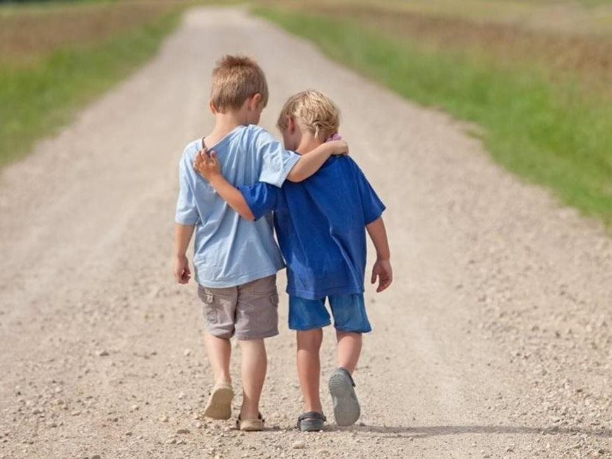Two children walking arm-in-arm down a gravel road between green fields