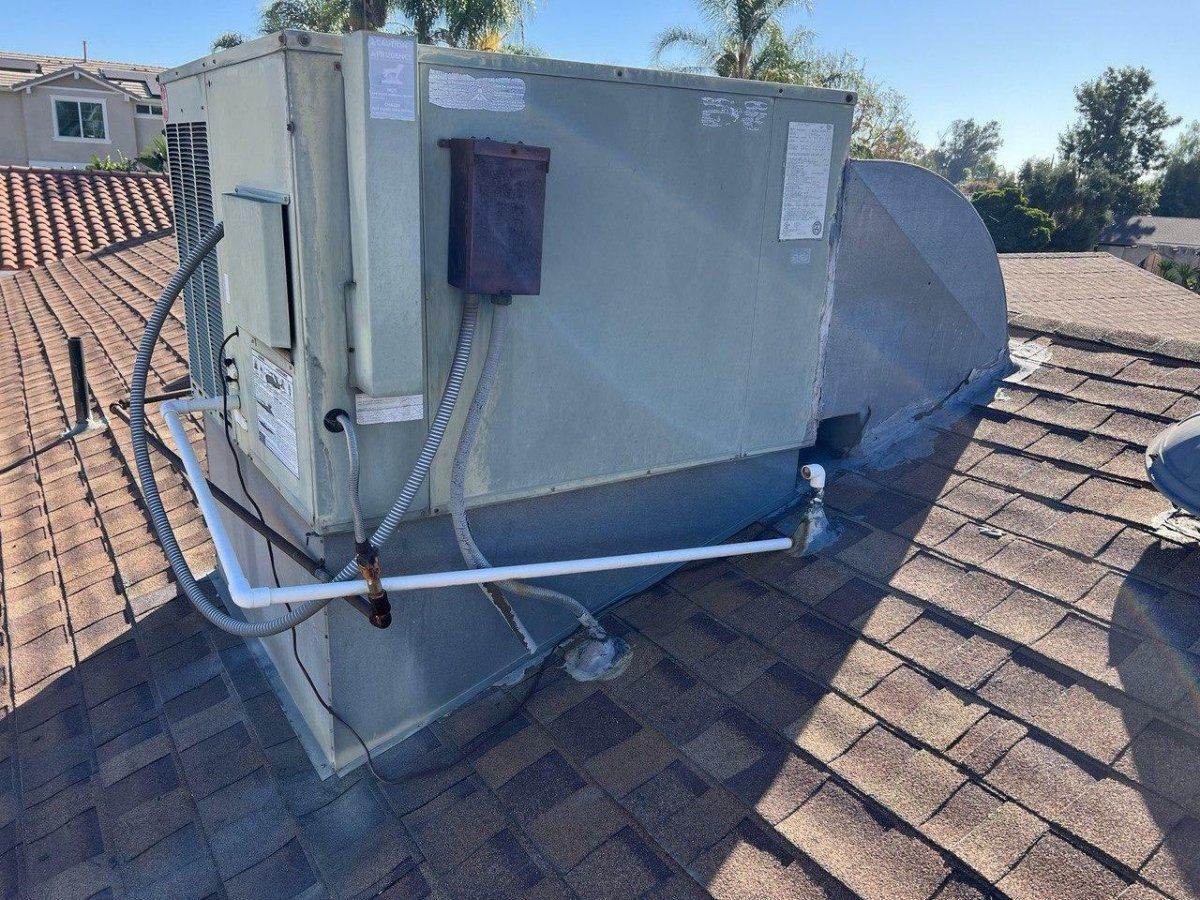 HVAC unit on a brown shingle roof, with a metal exterior and pipes visible. Bright sunny day.