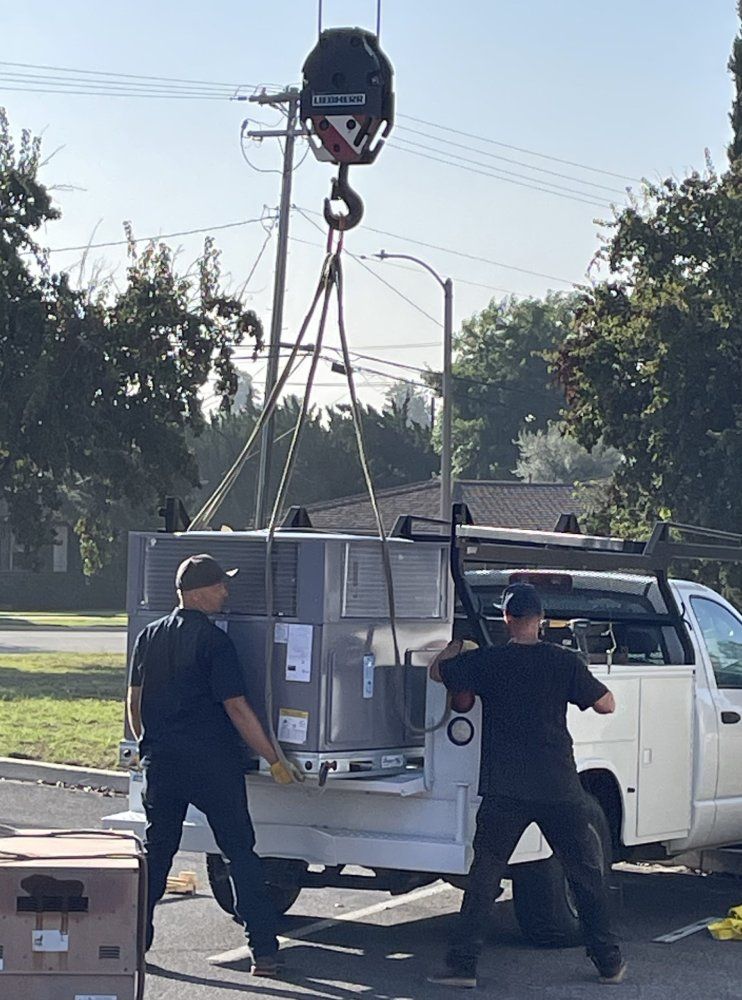 Two workers loading machinery from a truck with a crane. Sunny outdoors.
