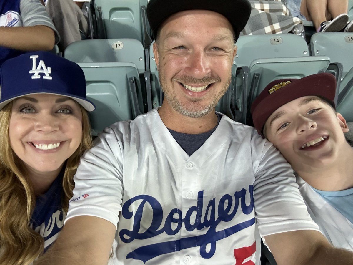 Family at a baseball game: man in Dodgers jersey, woman in a Dodgers hat, and a boy smiling in stadium seats.