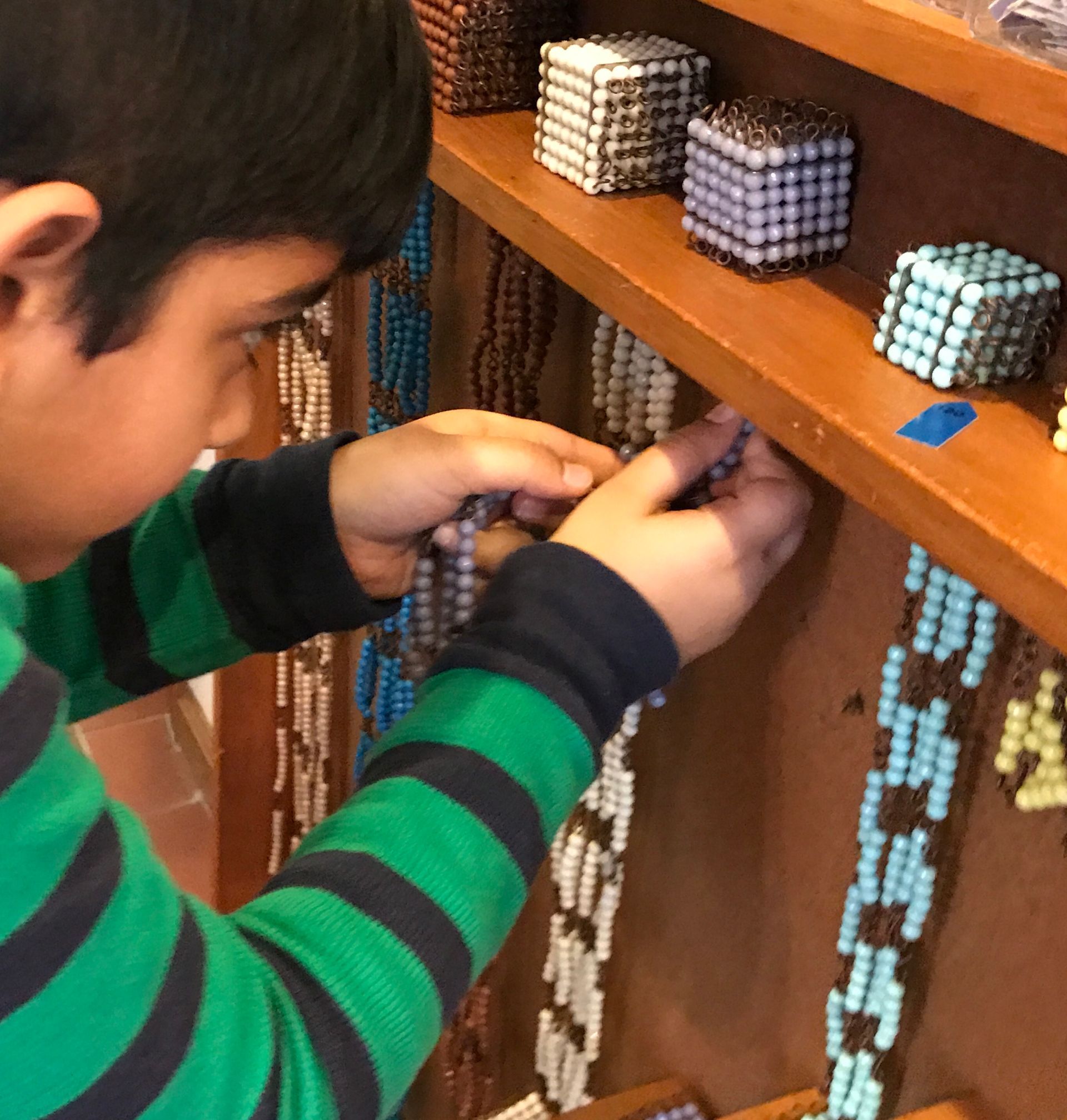A child in a striped green and black sweater arranges colorful beaded cubes on a wooden shelf with care and concentration.