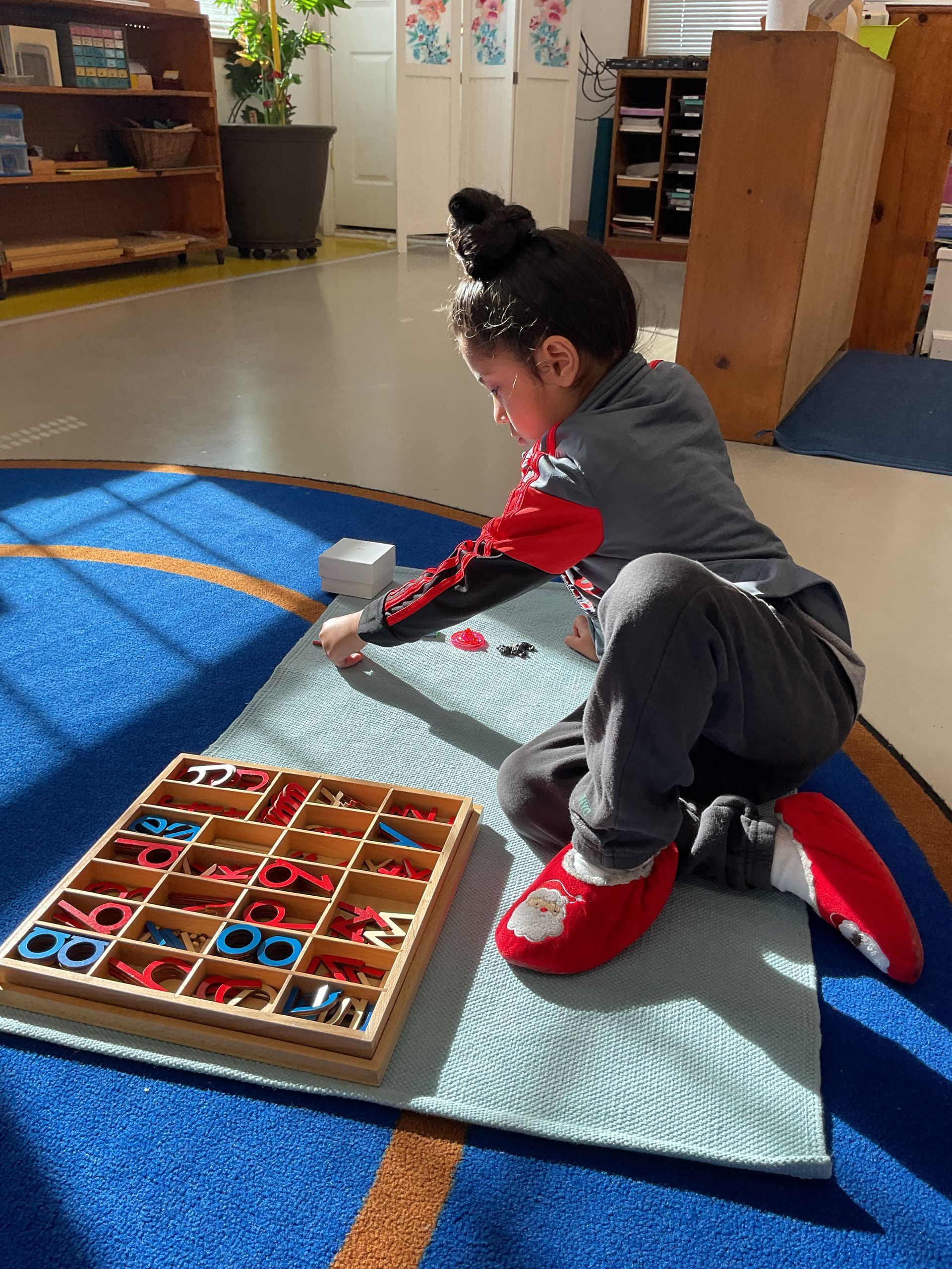 A child with a bun hairstyle sits on a blue rug, organizing colorful letters from a wooden box. Sunlight streams in the room.