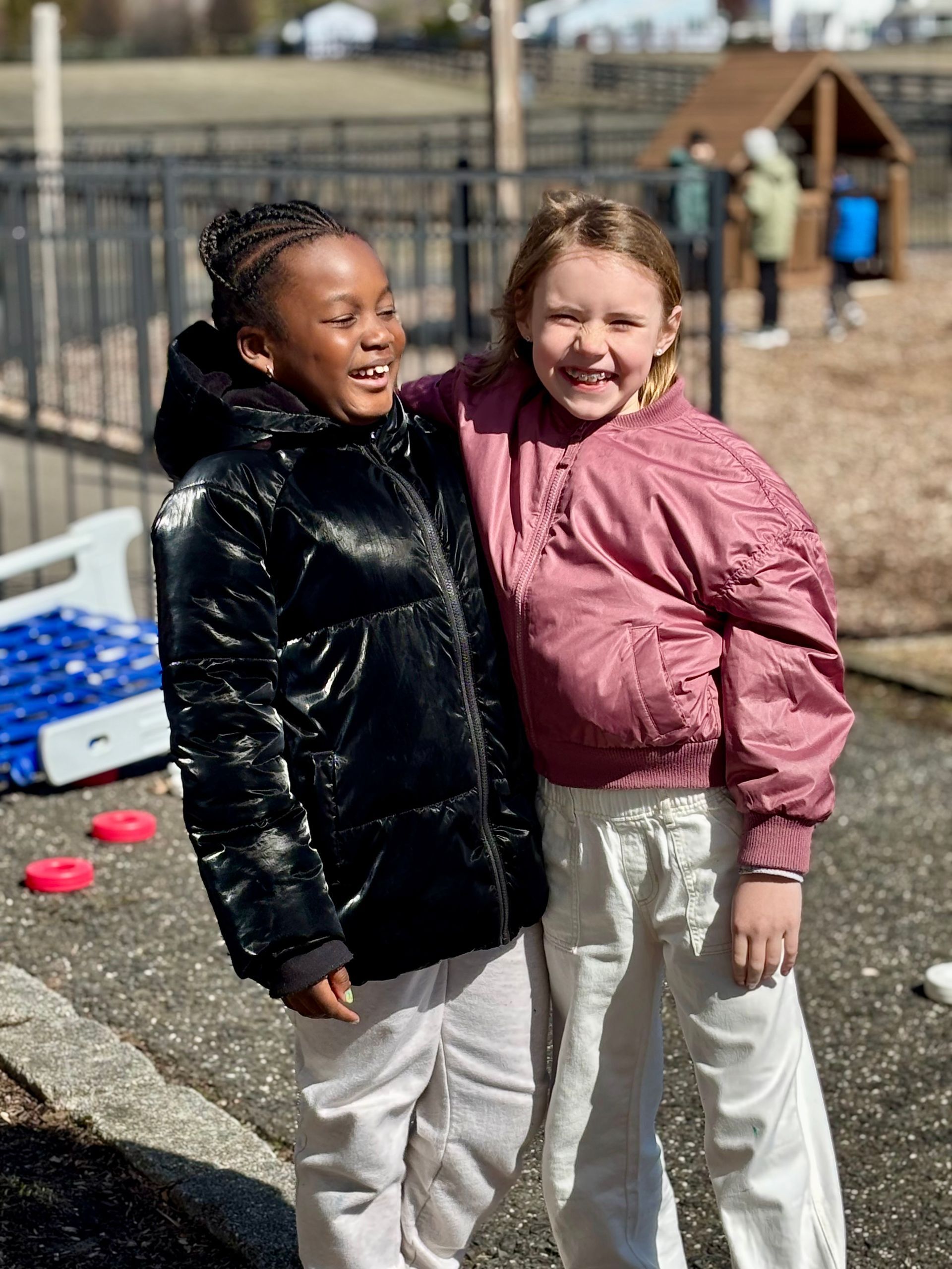 Two girls stand side by side outdoors, wearing stylish winter jackets, with a playground and kids playing in the background.