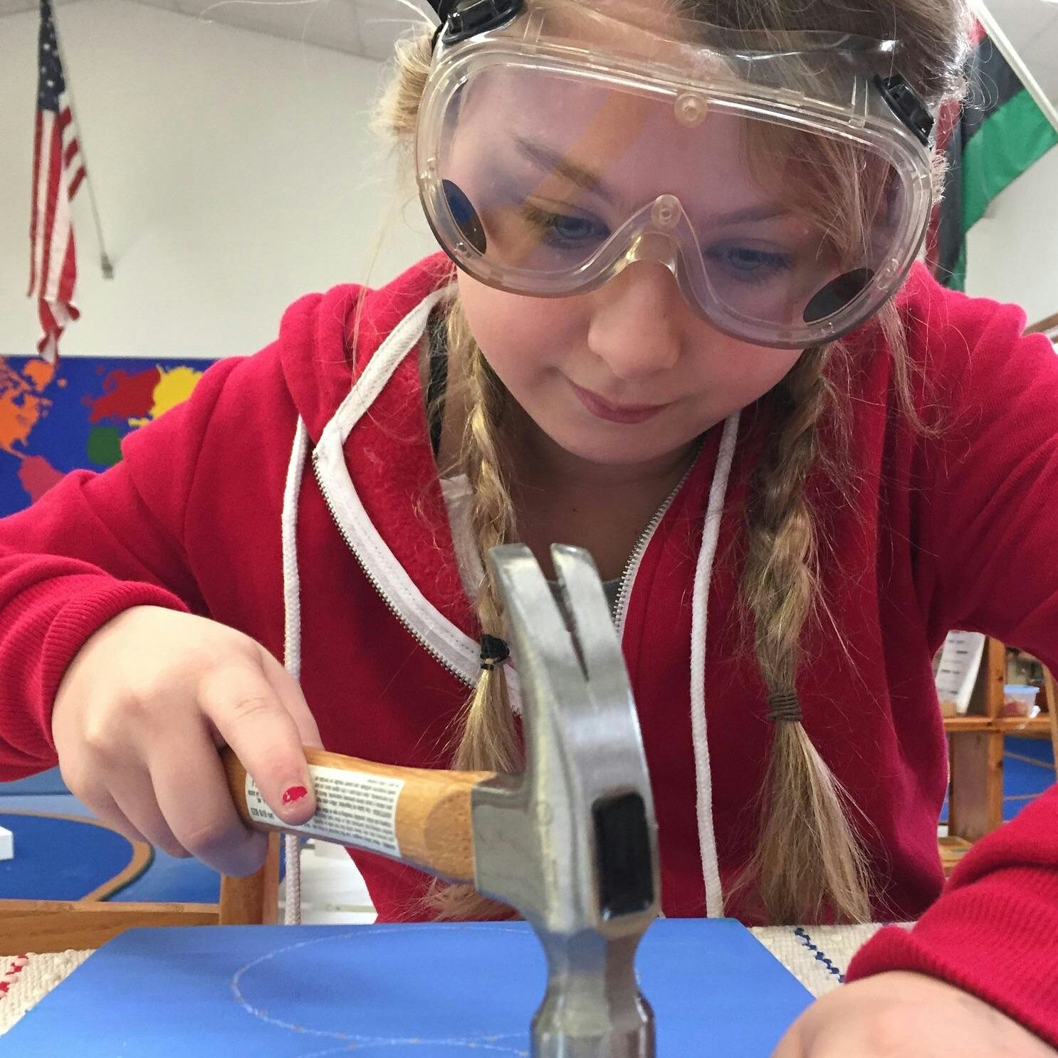 A young girl focused on using a hammer, wearing safety goggles and a red hoodie. Background features colorful world map.