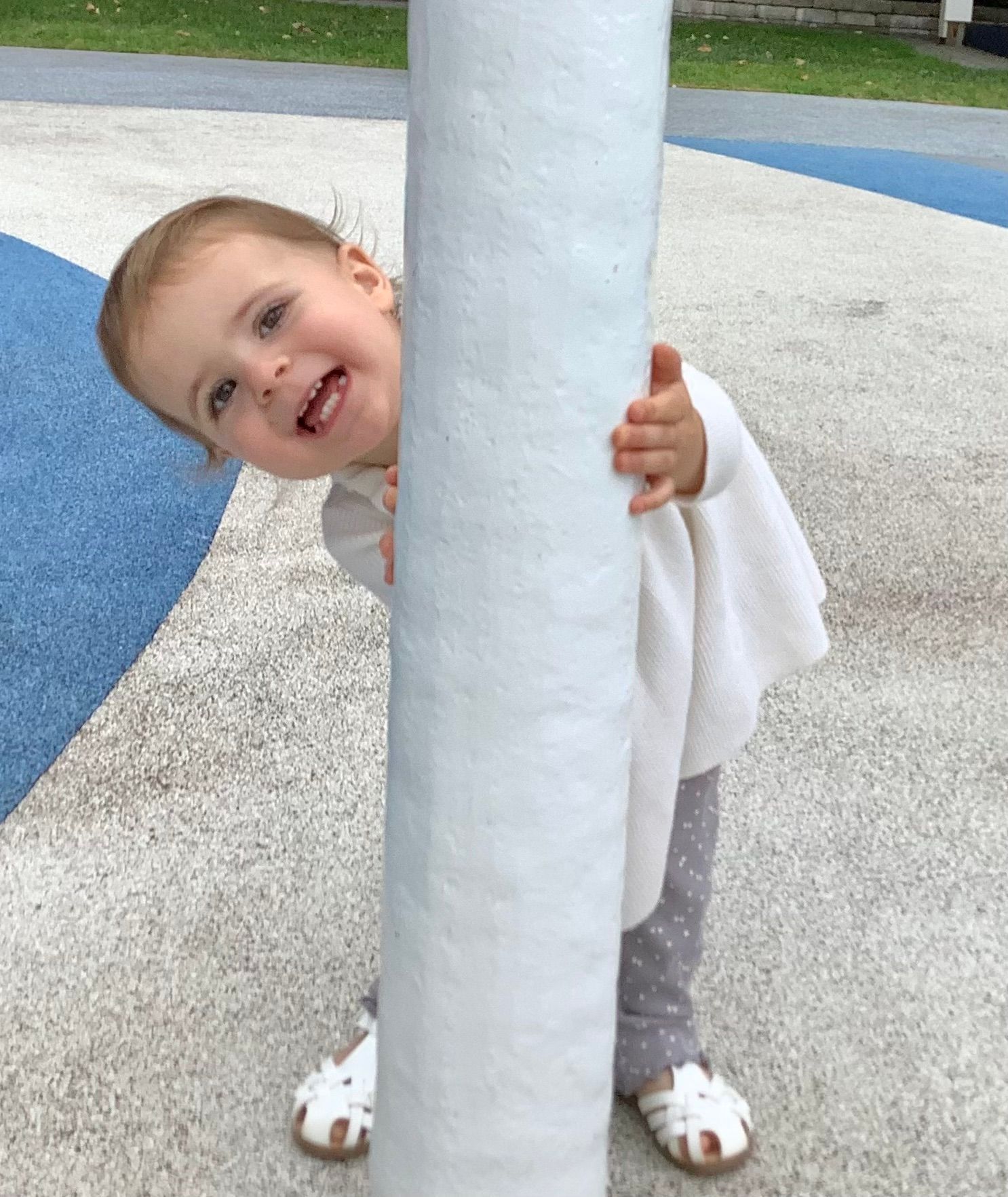 A child smiles brightly, playfully peeking from behind a white pole at a playground. The scene is joyful.