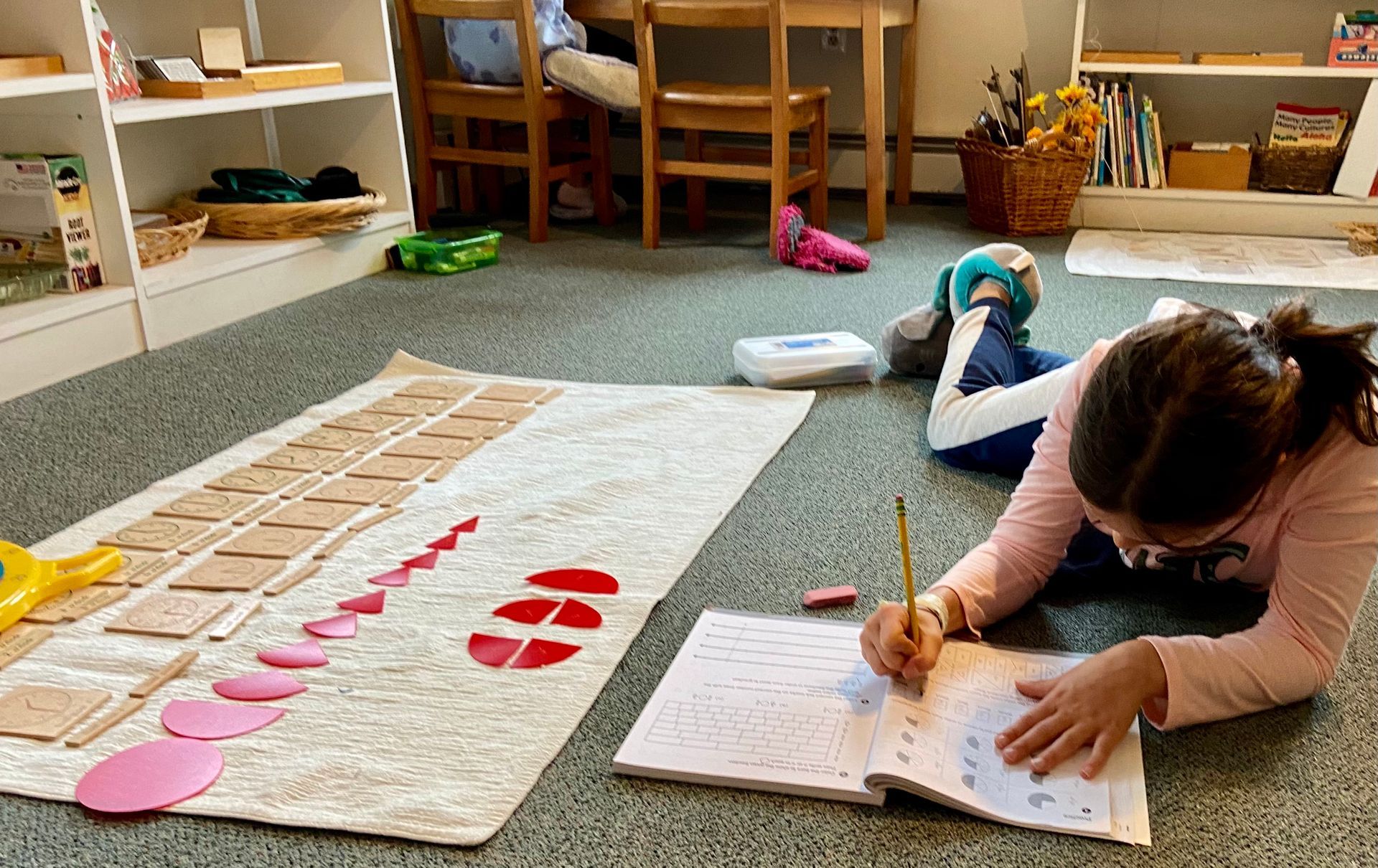 A child lies on a classroom floor, focused on writing in a workbook. Nearby, geometric shapes are arranged on a mat.