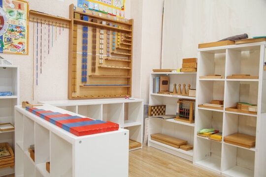 A brightly lit Montessori classroom features shelves with neatly organized educational materials, wooden blocks.