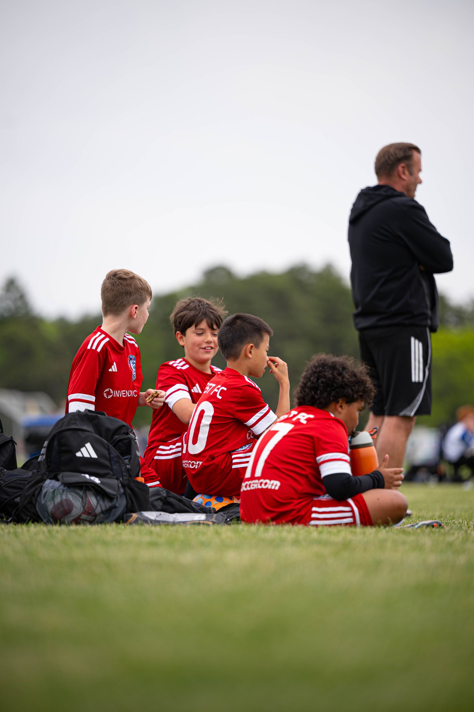 BEST FC soccer players in red jerseys sit on the sideline while a coach stands nearby on the field.