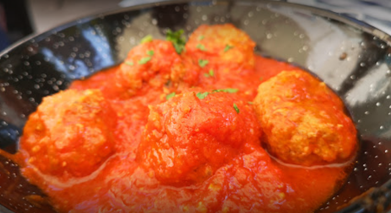Una ciotola bassa e scura, punteggiata di macchie, contenente diverse polpette dorate cotte a fuoco lento in una salsa di pomodoro rosso acceso.