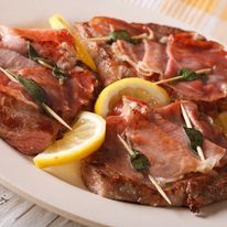 Cotolette di vitello saltimbocca guarnite con prosciutto e salvia, fissate con stuzzicadenti e servite su un piatto con fette di limone.
