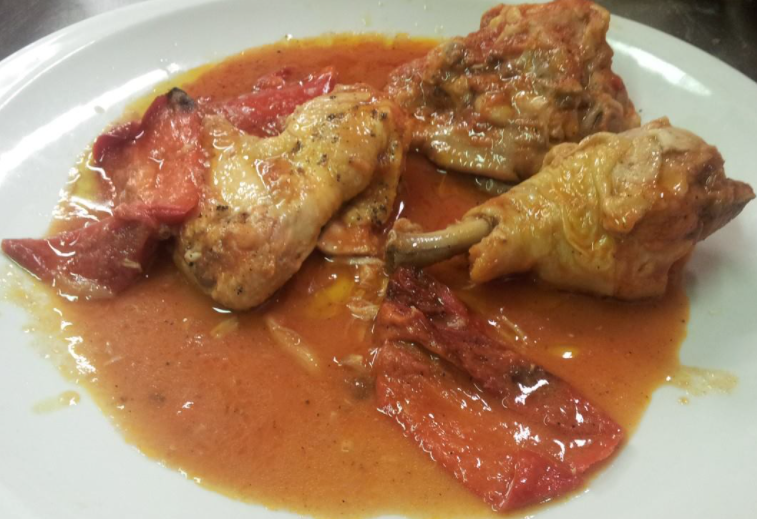 Una porzione di pezzi di pollo brasati in una salsa rossastra a base di pomodoro, con grossi pezzi di peperoni rossi arrostiti su un piatto.