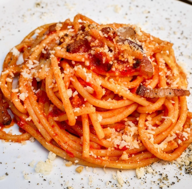 Primo piano di un piatto di bucatini al sugo di pomodoro, guarniti con formaggio grattugiato e pezzetti di lardo stagionato.