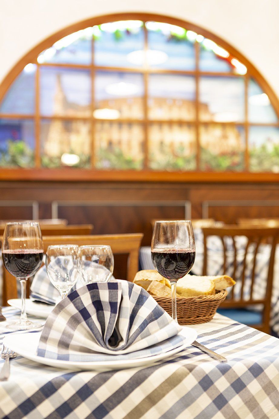 Un tavolo di ristorante apparecchiato con due bicchieri di vino rosso, un tovagliolo e un cestino di pane, di fronte a una suggestiva finestra ad arco.