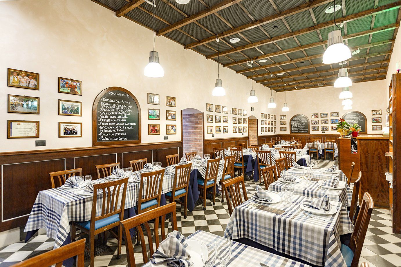 L'interno di un ristorante ben illuminato presenta diversi tavoli con tovaglie a quadri, sedie in legno e decorazioni incorniciate.