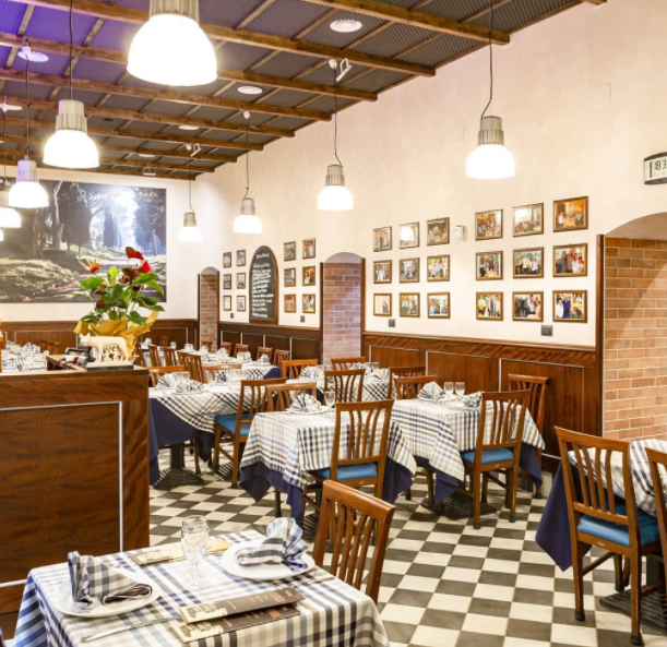 Sala da pranzo con pavimento a scacchi, tavoli con tovaglie blu e bianche, lampade a sospensione e foto incorniciate alle pareti.