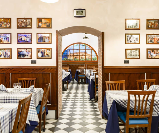 Interno di un ristorante con pavimento a scacchi, tavoli con tovaglie blu e bianche e quadri incorniciati alle pareti.