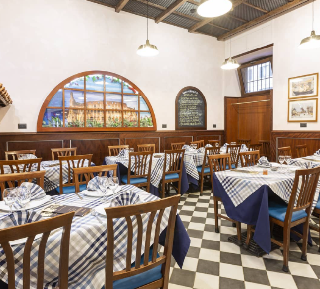 La sala da pranzo di un ristorante con tovaglie a quadri bianchi e blu, sedie di legno e una finestra che raffigura un edificio.