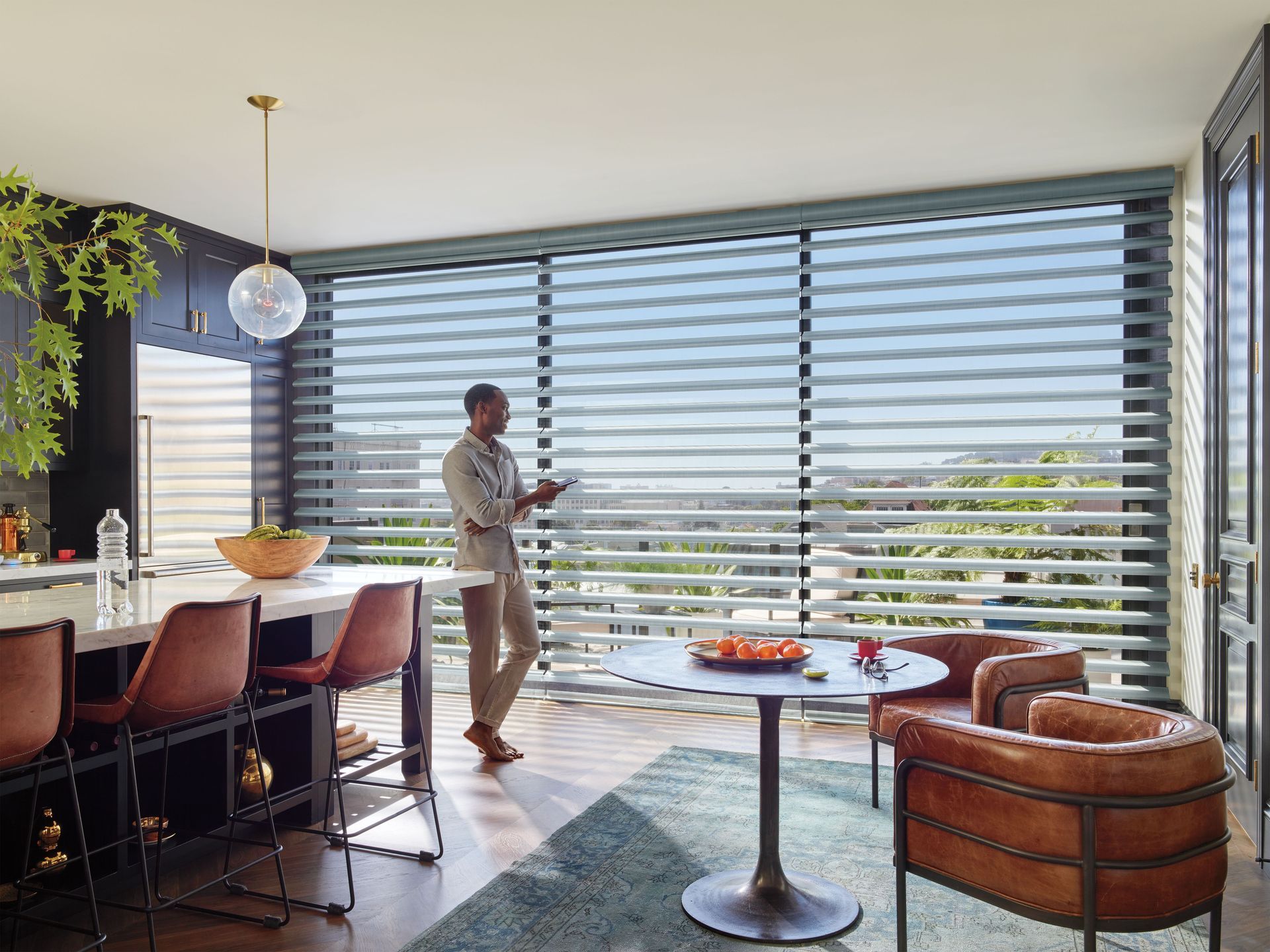 Man operating automated blinds in a modern kitchen, overlooking a cityscape.