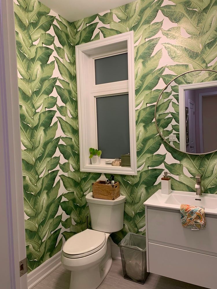 Bathroom with white toilet, gray vanity, and green leaf wallpaper. A round mirror hangs above the sink.