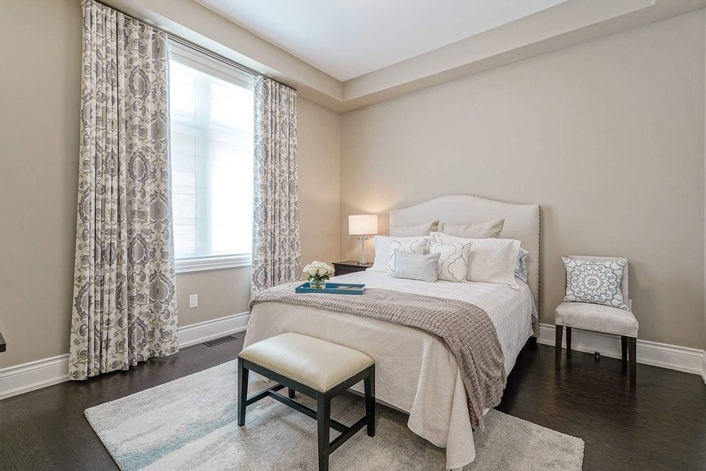 Bedroom with beige walls, dark wood floor, bed with white bedding, patterned curtains, and a bench.