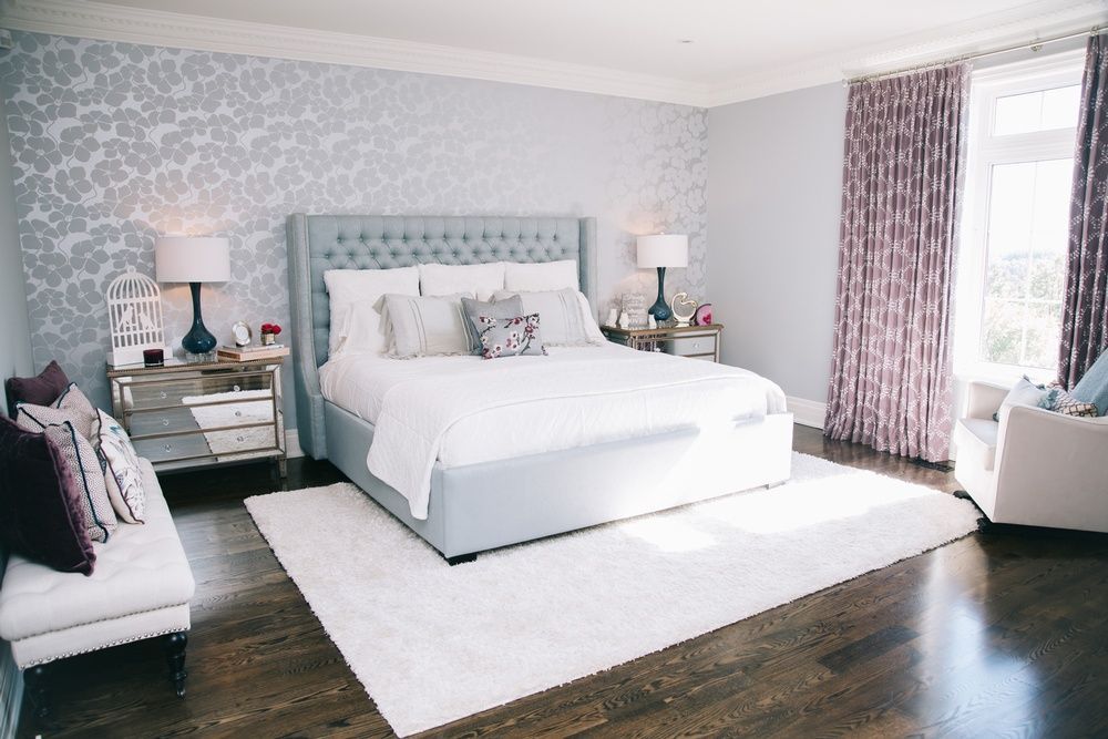 Bedroom with light blue upholstered bed, matching bedside tables, floral wallpaper, purple curtains, and a white rug.
