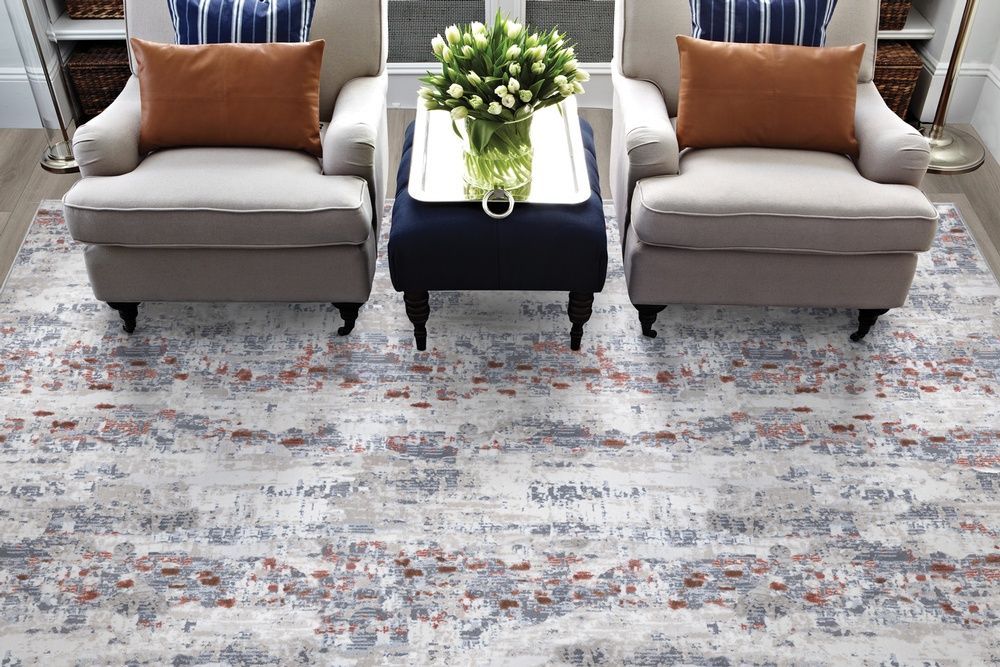 Two armchairs with orange pillows face each other, with a blue ottoman and rug with a floral pattern.