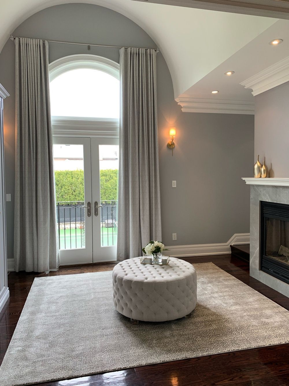Living room with arched window, light gray walls, fireplace, and tufted ottoman.